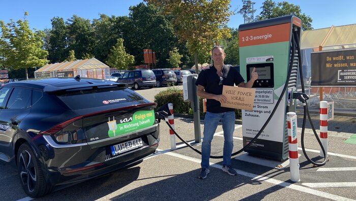 Elektroauto von Pfalzwerke lädt an einer öffentlichen Ladesäule; daneben steht eine Person mit Schild ‚Volle Power voraus‘ auf einem Parkplatz im Freien.