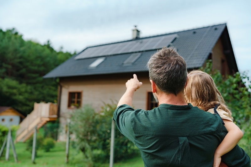 Vater mit Kind auf dem Arm zeigt in Richtung eines Hauses mit PV-Anlage auf dem Dach 