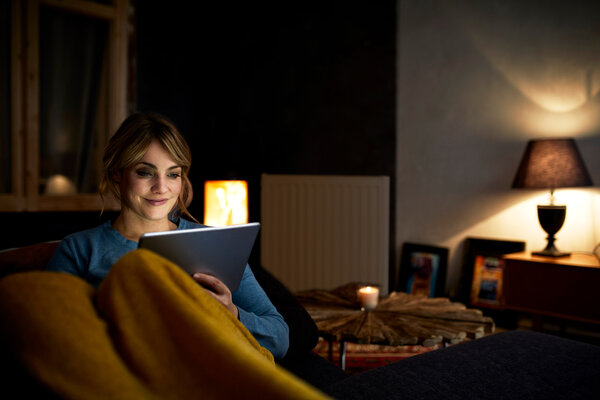 Das Bild zeigt eine Frau, die abends bei gedämpftem Licht  mit einem Tablet auf der Couch sitzt.  | © Pfalzwerke VIDA macht das Zuhause smart – auf der ImmobilienMesse
