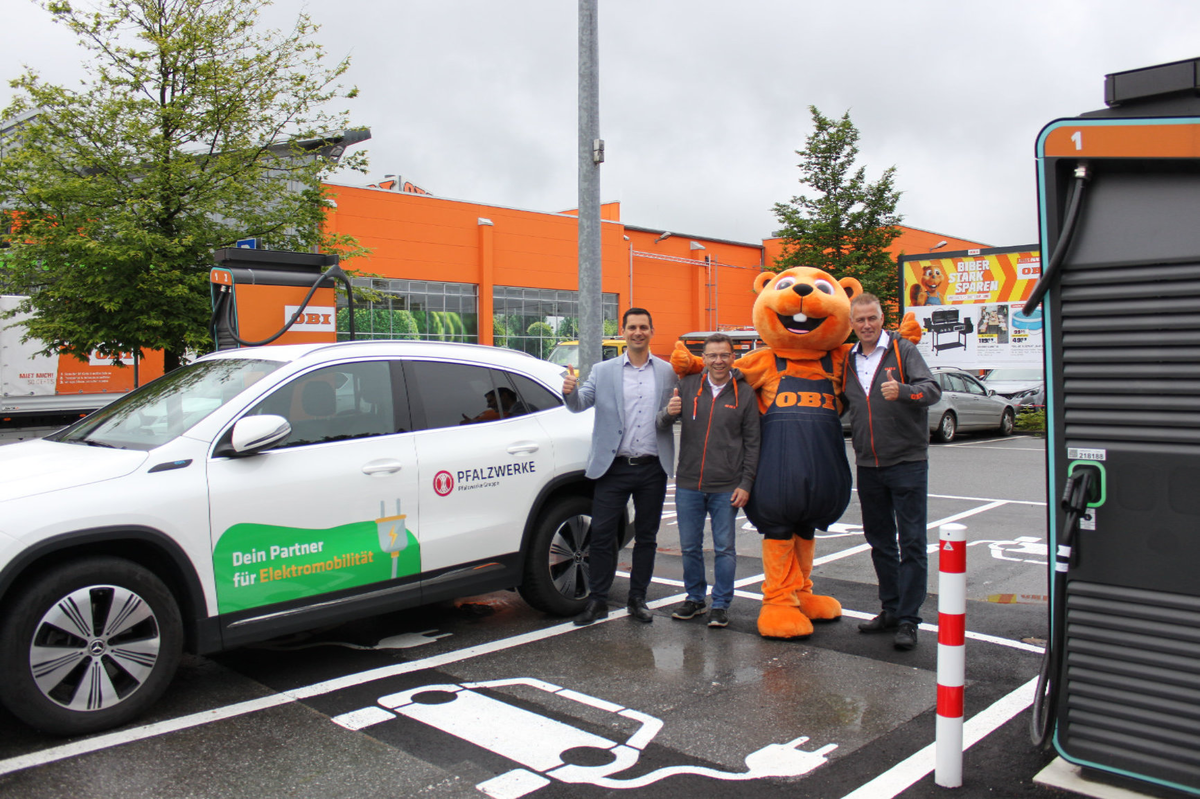 Drei Menschen stehen an der Ladesäule auf dem OBI Parkplatz und strecken den Daumen in die Höhe. 