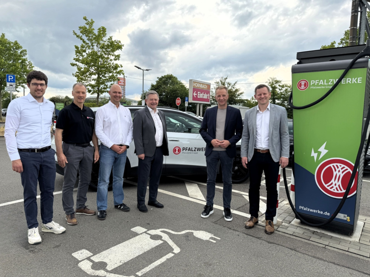 Mehrere Personen stehen an einem Pfalzwerke-Elektrofahrzeug neben einer E-Ladesäule auf dem Parkplatz eines Hornbach-Markts.